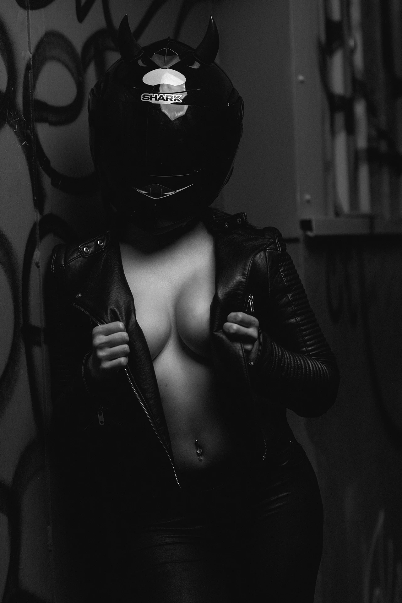 shoot-charme-02 Une silhouette féminine énigmatique, casquée et vêtue de cuir, évolue dans un décor urbain sombre, entre ombre et lumière.
