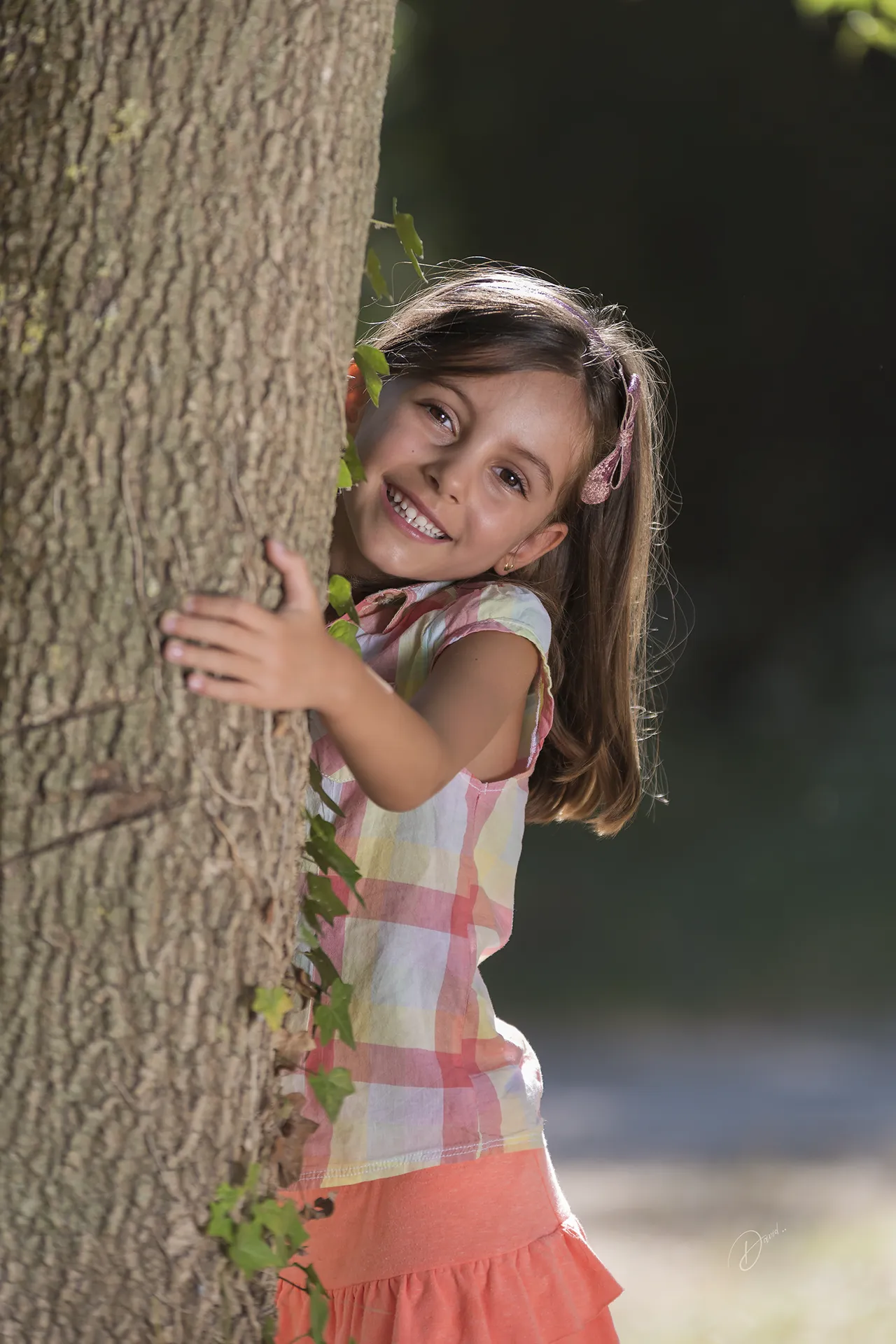 Enfant souriante cachée derrière un arbre lors d’un shooting enfant Côte d’Azur en extérieur.