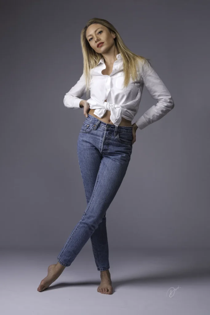 Portrait mode en studio, femme en chemise blanche nouée et jean bleu, pose déhanchée sur fond gris uni