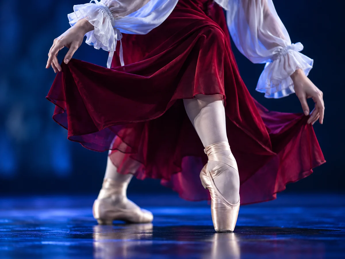 Gros plan sur les pointes d'une danseuse et sa jupe bordeaux en mouvement, sous une lumière bleue.